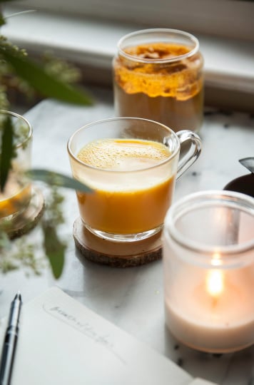 A head on shot of a creamy orange liquid in a clear mug. A lit candle and jar of turmeric are nearby.