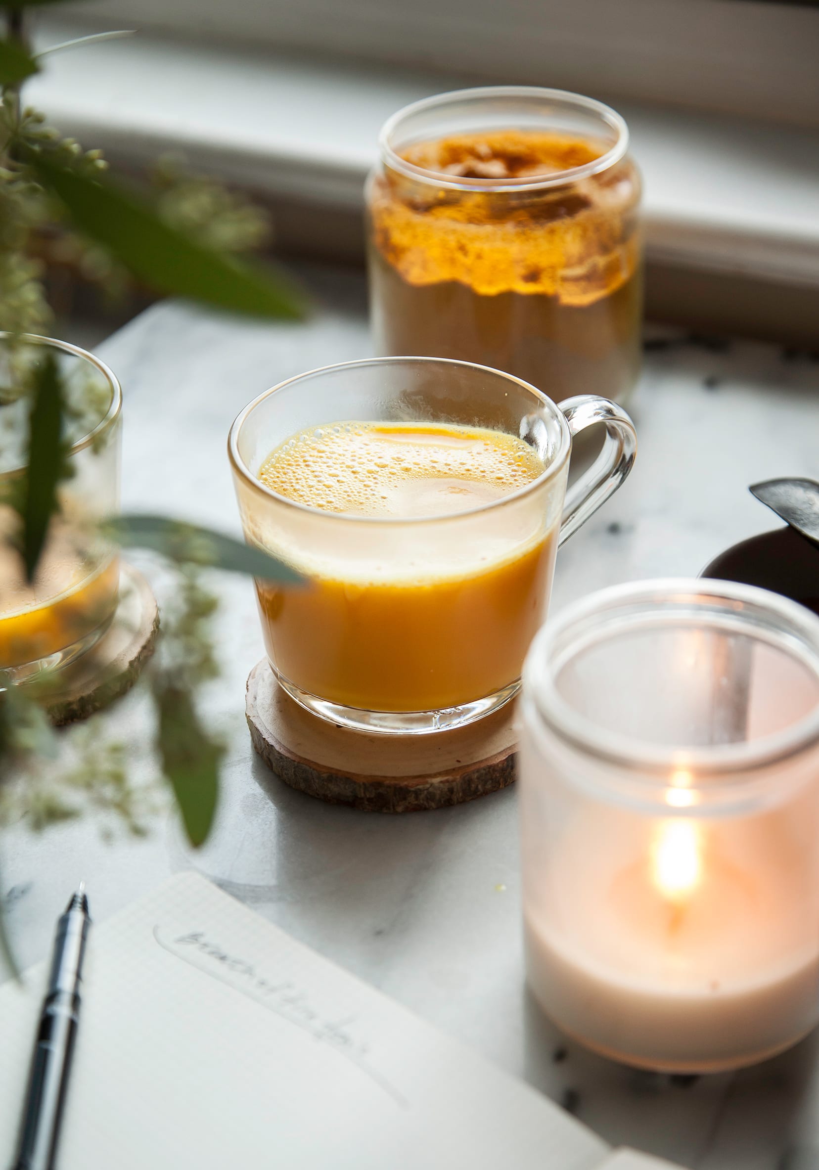 A head on shot of a creamy orange liquid in a clear mug. A lit candle and jar of turmeric are nearby.