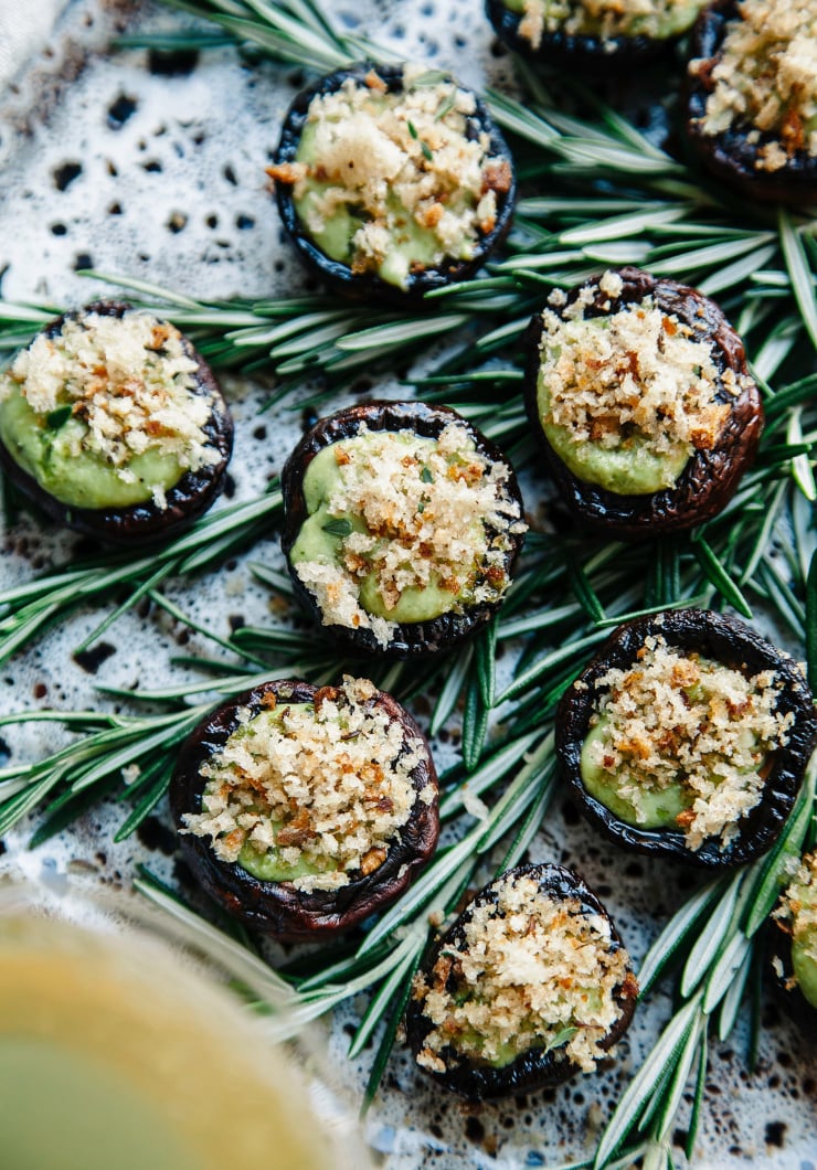 An overhead shot of stuffed cremini mushrooms on a stoneware plate. The mushrooms have a pale green creamy sauce inside the caps and a topping of golden brown breadcrumbs.