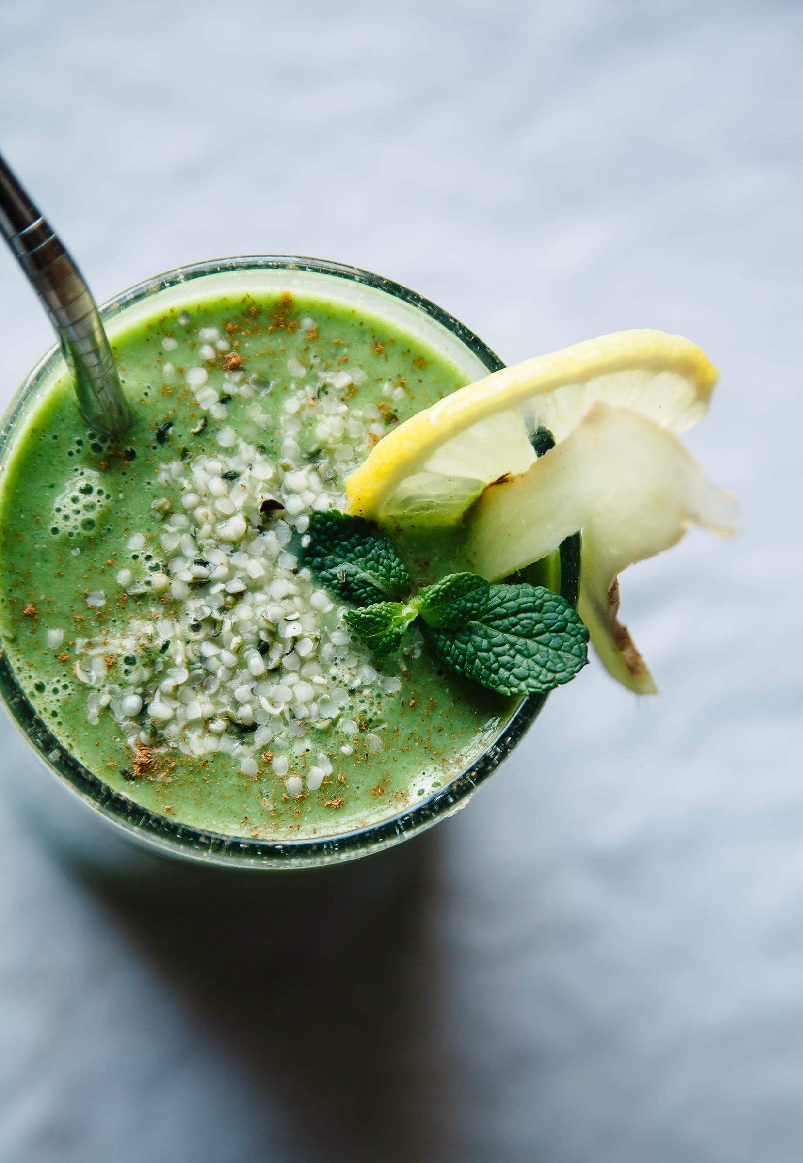 An up close, overhead shot of a green smoothie in a glass. The top of the smoothie is sprinkled with hemp seeds and a sprig of mint. A slice of lemon is tucked into the rim of the glass.