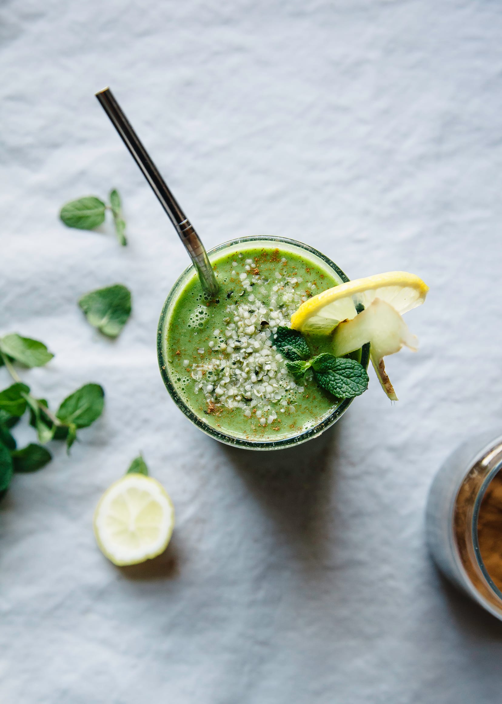 An overhead shot of a green smoothie in a glass. The top of the smoothie is sprinkled with hemp seeds and a sprig of mint. A slice of lemon is tucked into the rim of the glass.