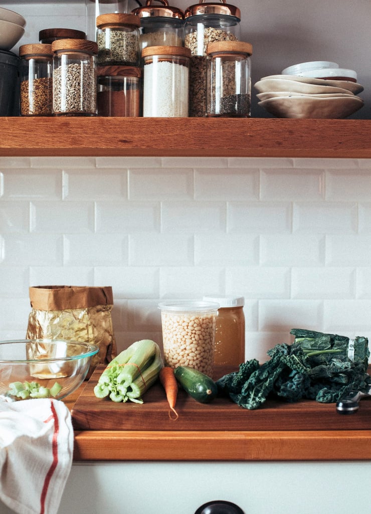 A shot of my kitchen’s open shelving and countertop. On the counter: a container of white beans, vegetable stock, chopped kale, a carrot, celery, and a dish towel.