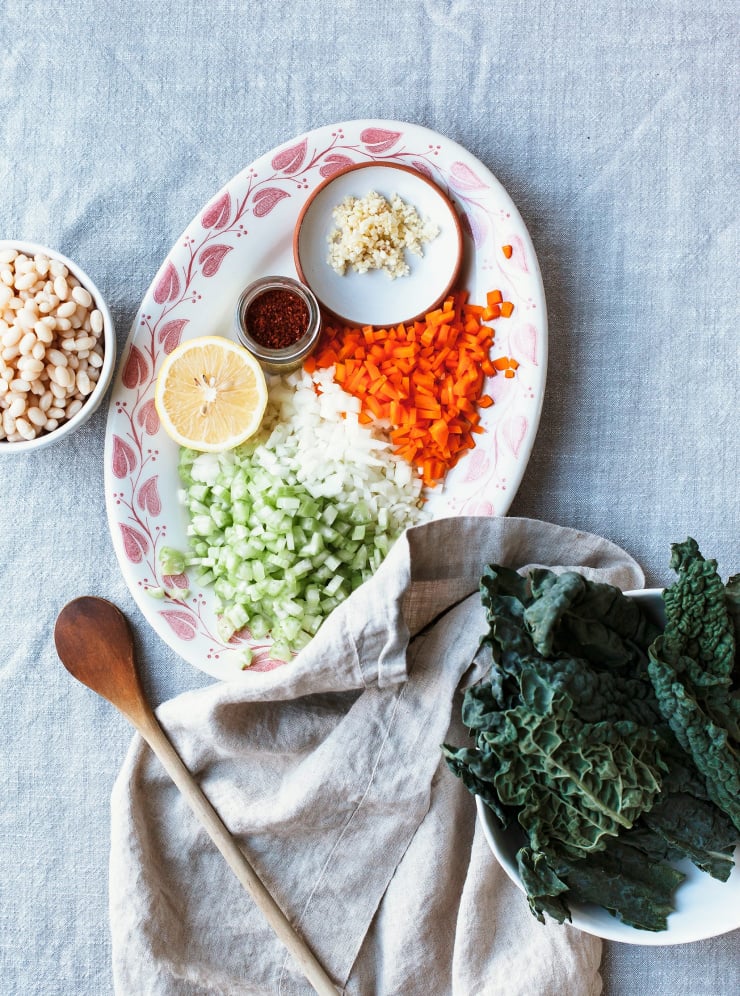 Chopped prep for making soup. On a white platter with red leaf detail on the edges: chopped celery, onions, carrots, garlic, chili flakes, and a lemon half. To the side, there’s chopped kale and white beans. 