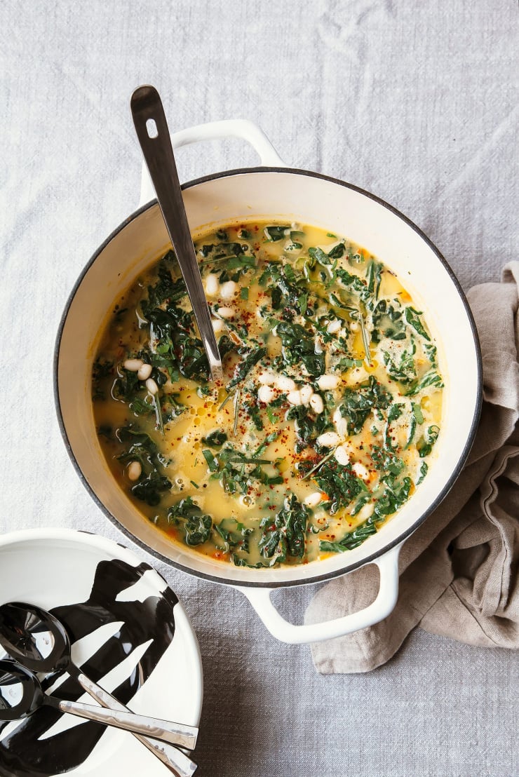 An overhead shot of a pot of creamy white bean soup with kale, rosemary and lemon. The pot os off white with bowls and spoons to the side, over a grey linen tablecloth.