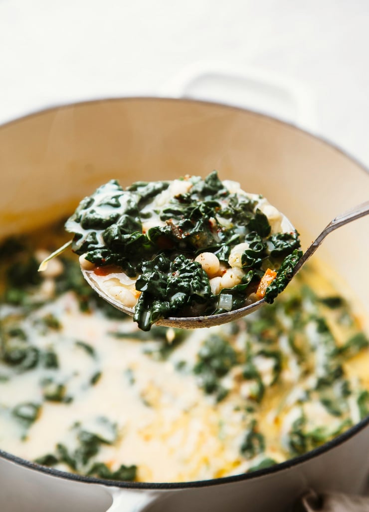 A 3/4 angle shot of a ladle of white bean soup with kale, emerging from an off-white pot.