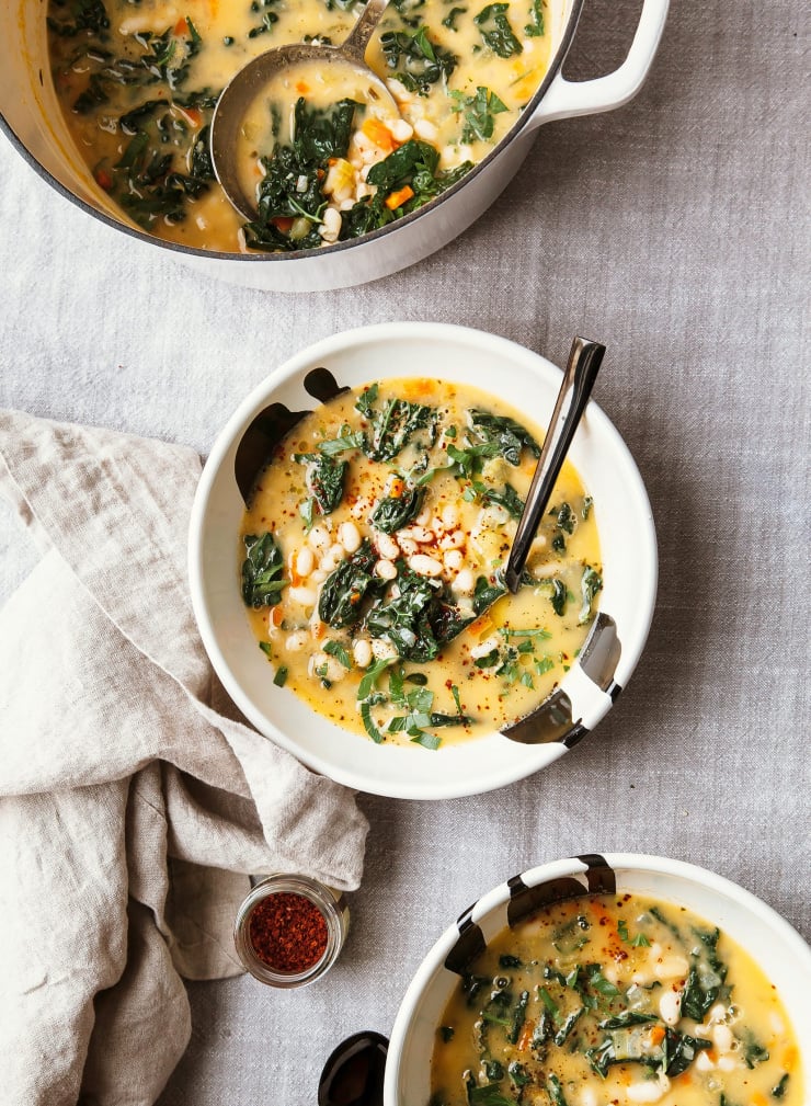 An overhead shot of 2 bowls of creamy white bean soup, with the big pot of it to the side. There is a beige table linen rumpled nearby and everything is set over a grey linen tablecloth.