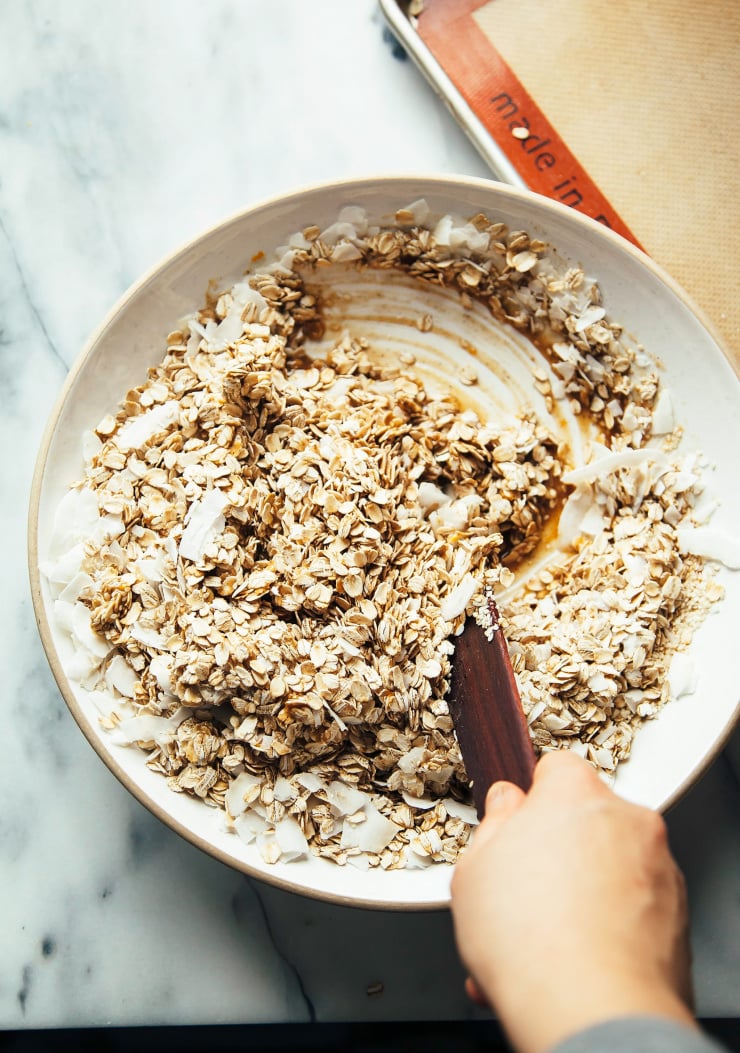 Overhead image shows a hand stirring together a granola mixture, pre-baking.