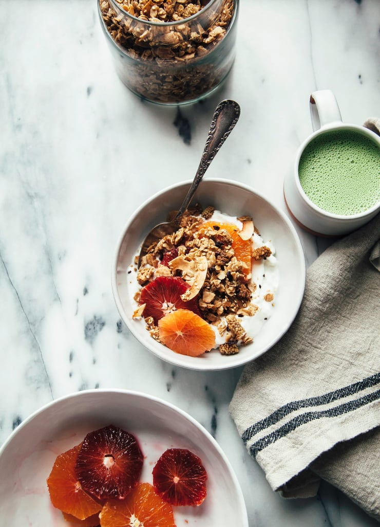 An overhead shot of tahini granola in a bowl with yogurt and citrus. Cut citrus, a light green matcha latte, and a jar of granola are seen to the side.