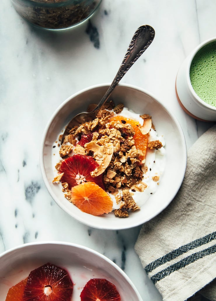An overhead shot of tahini granola in a bowl with yogurt and citrus. Cut citrus, a light green matcha latte, and a jar of granola are seen to the side.