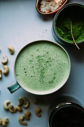 An overhead shot of a mellow green matcha latte with a bowl of matcha nearby and some cashews strewn about.