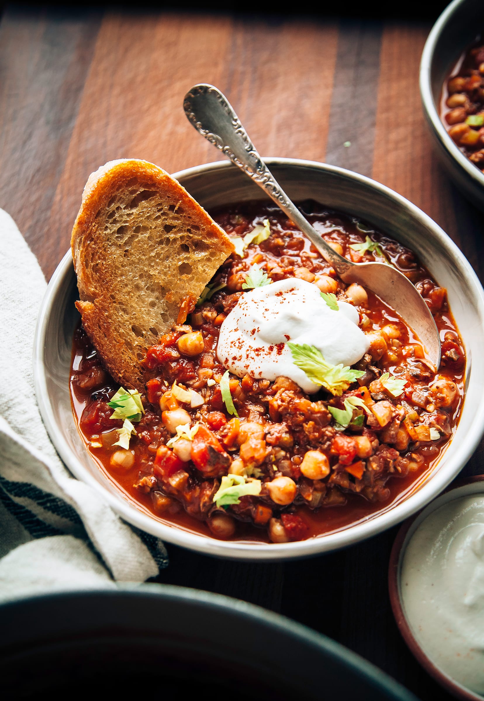 An slight 3/4 angle shot of Buffalo chickpea chili in a wide bowl with a piece of toasted bread on top. There’s also a bright white dollop of cashew four cream.