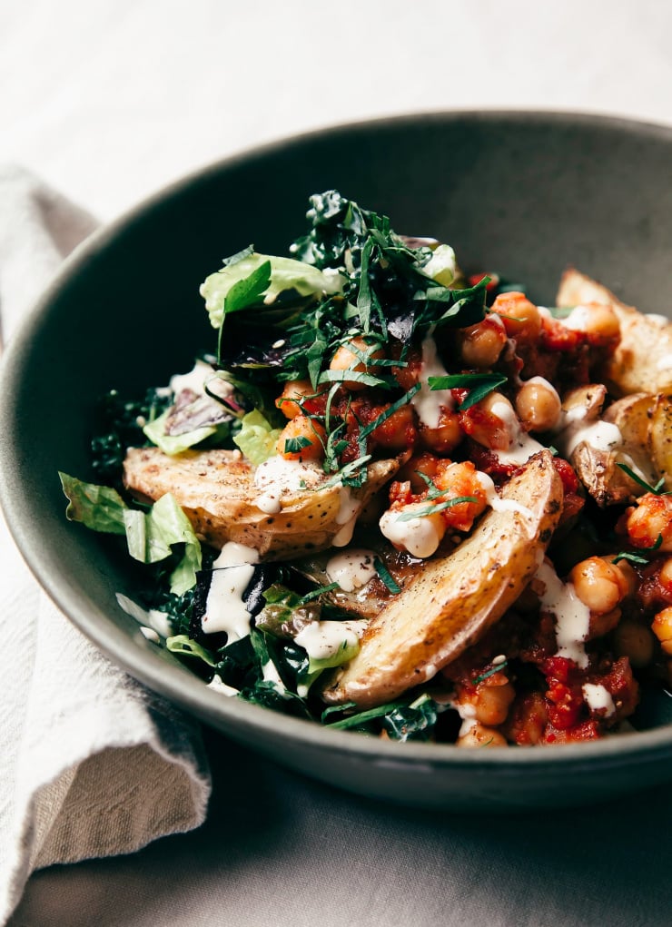 A 3/4 angle shot of a dark green bowl filled with chickpeas in a red sauce, wedges of roasted potatoes, chopped greens, and a creamy, white sauce drizzled on top.