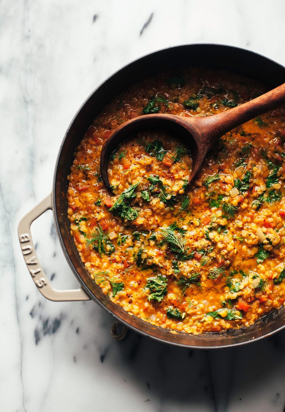 An overhead shot of quick smoky red lentil stew in a beige, cast iron braiser-style pot. There is a wooden spoon sticking out of the orange stew. The stew is garnished with chopped parsley.