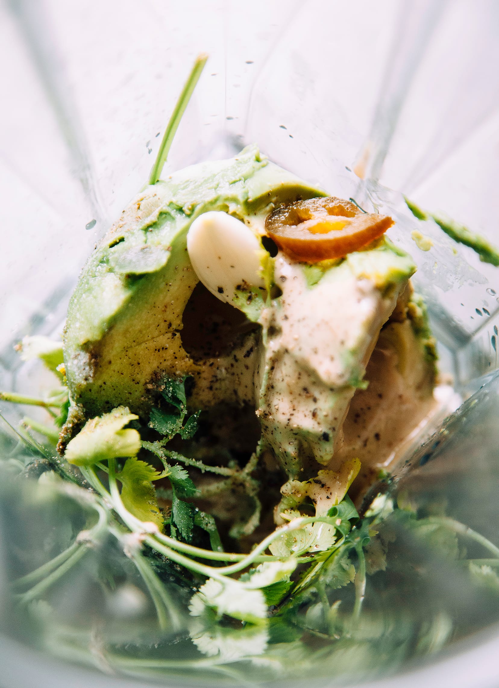 Overhead shot of the contents of a blender pitcher: avocado, cilantro, tahini, jalapeno, garlic, salt, and pepper.