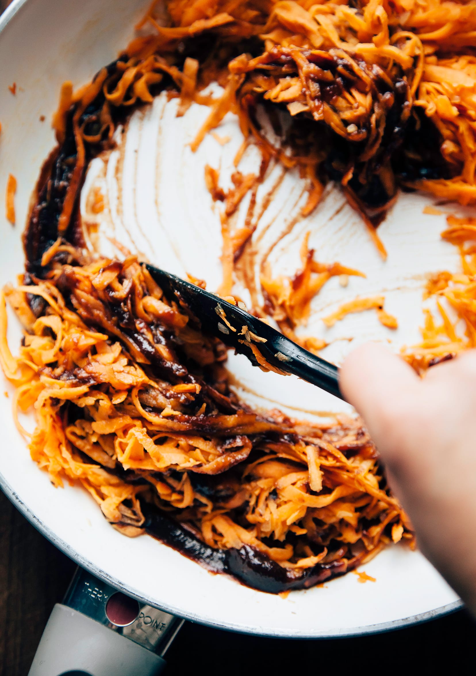 Image shows a hand stirring shredded sweet potatoes and BBQ sauce together in a sauté pan.