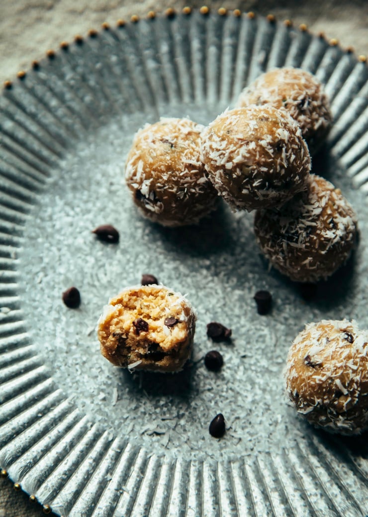 Image shows rolled oatmeal cookie bites, coated in shredded coconut on a metal plate. A bite is taken out of one, revealing chocolate chips.