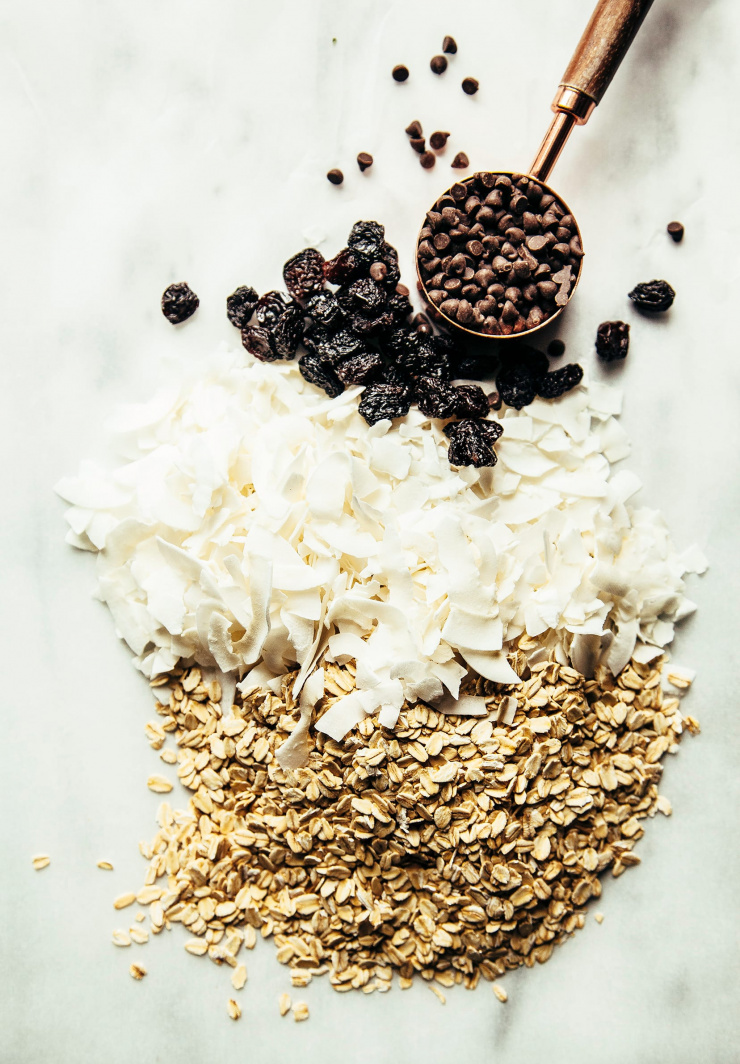 An overhead shot of chocolate chips, coconut flakes, raisins, and rolled oats, all lined up together.
