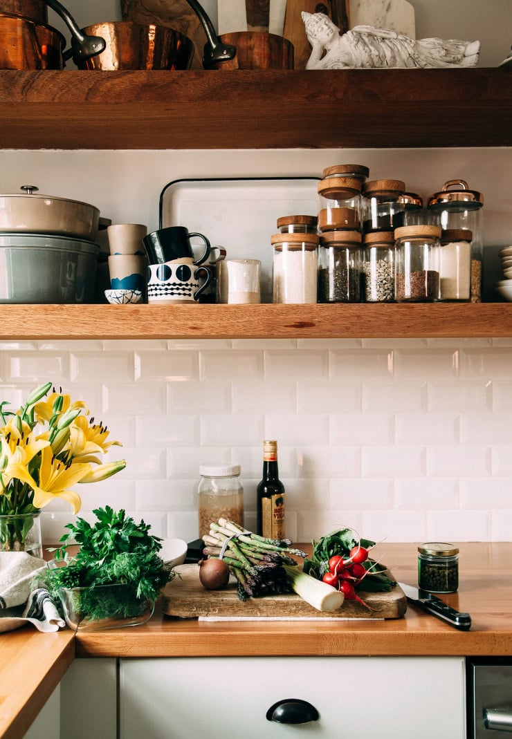 Image shows a kitchen scene with ingredients and cut lilies on the counter. The kitchen has open wood shelves and shiny white subway tile in the background.