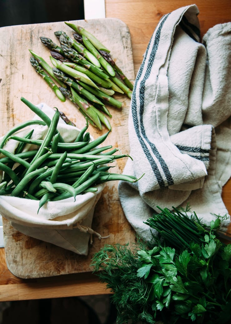 Image shows a wooden cutting board with asparagus, green beans, and herbs nearby or on top. A beige linen towel is nearby as well.