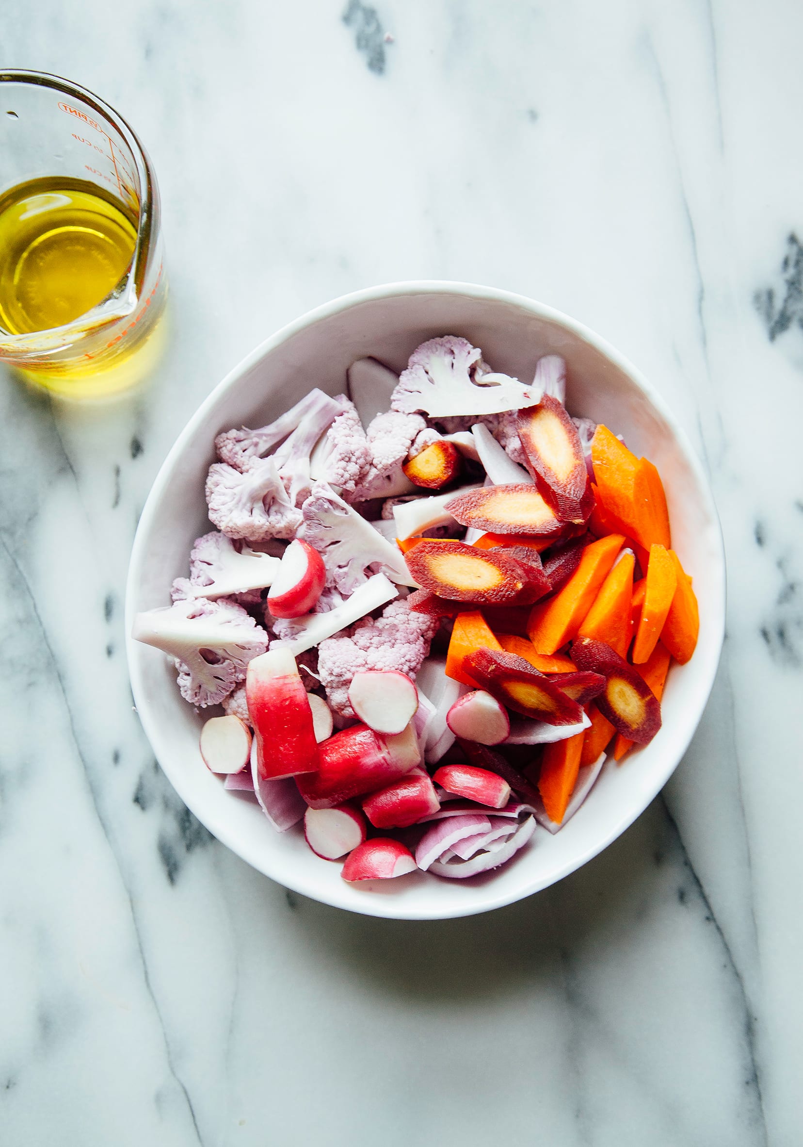 An overhead shot of cut vegetables in a white bowl: purple cauliflower, carrots, radishes, and onions.