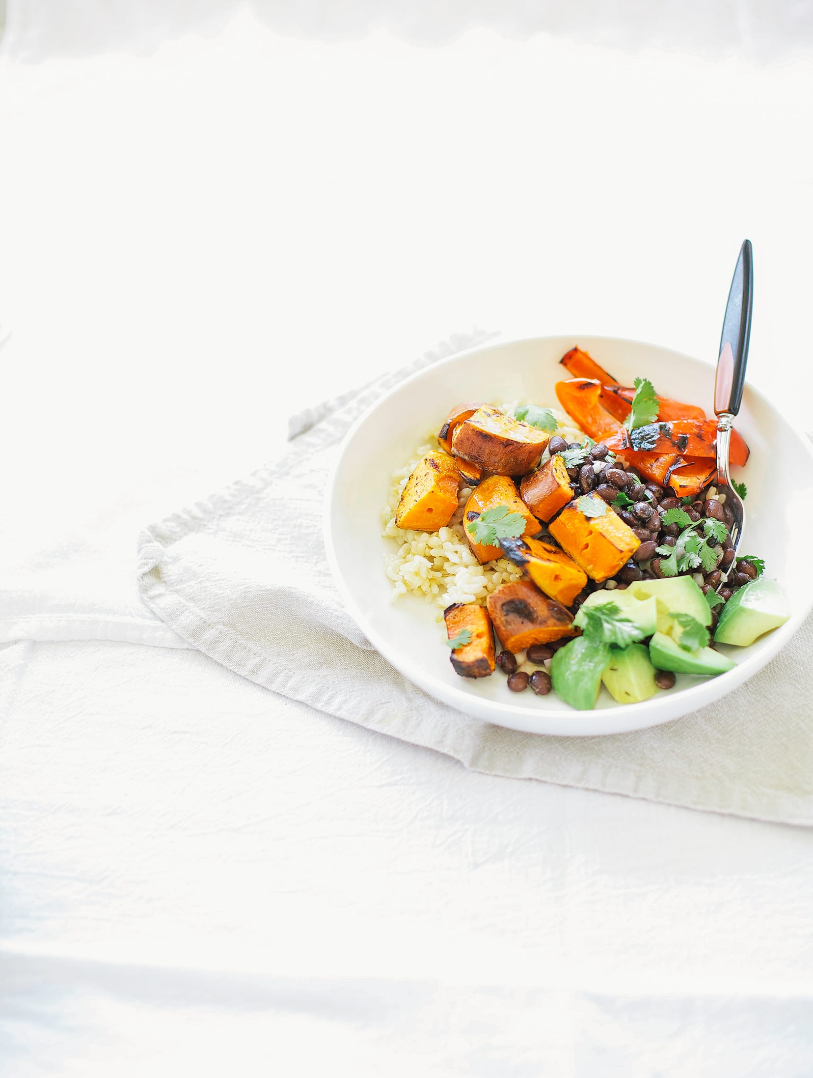 A 3/4 angle shot of a white bowl on a white background filled with brown rice, black beans, grilled sweet potatoes, cooked red bell pepper pieces and chopped avocado