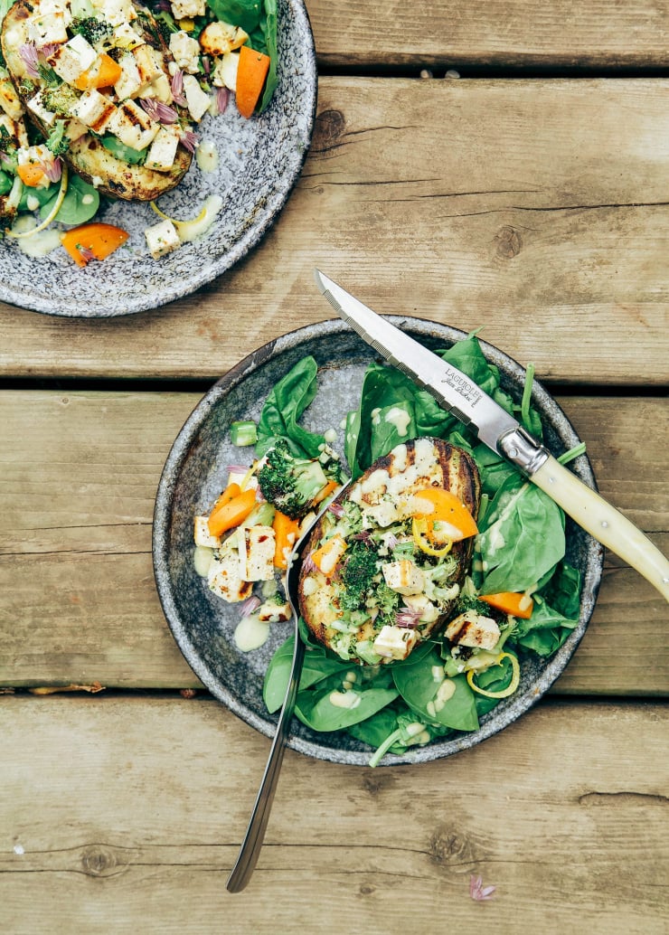 An overhead shot of a grilled avocado half stuffed with a cooked tofu, apricot, and broccoli mixture. The dish is served with some baby spinach and a creamy sauce. Part of a roundup of vegan tofu recipes.