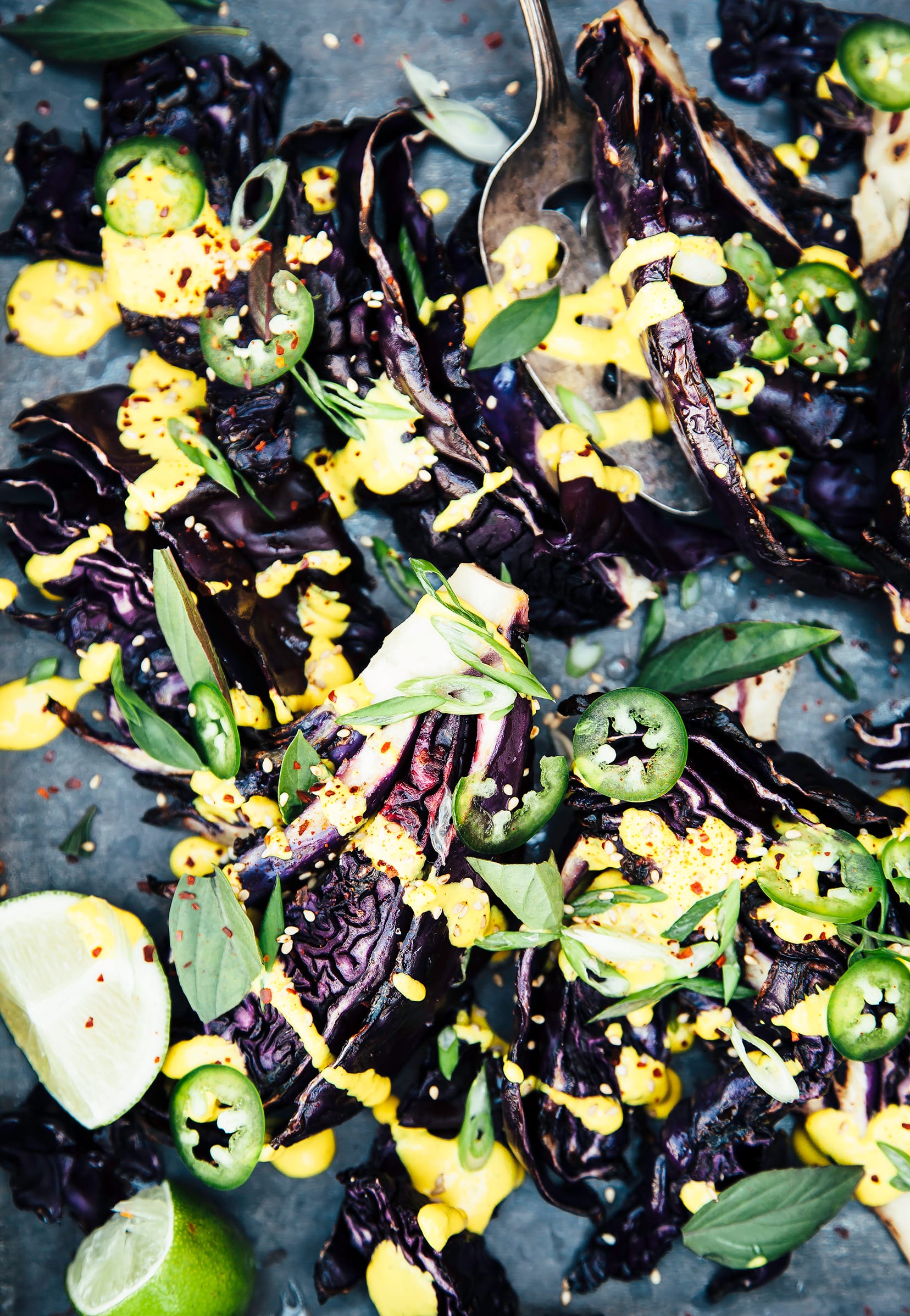 An up close and overhead shot of grilled purple cabbage wedges, topped with drizzles of a creamy yellow sauce, slices of jalapeño, and chopped herbs.