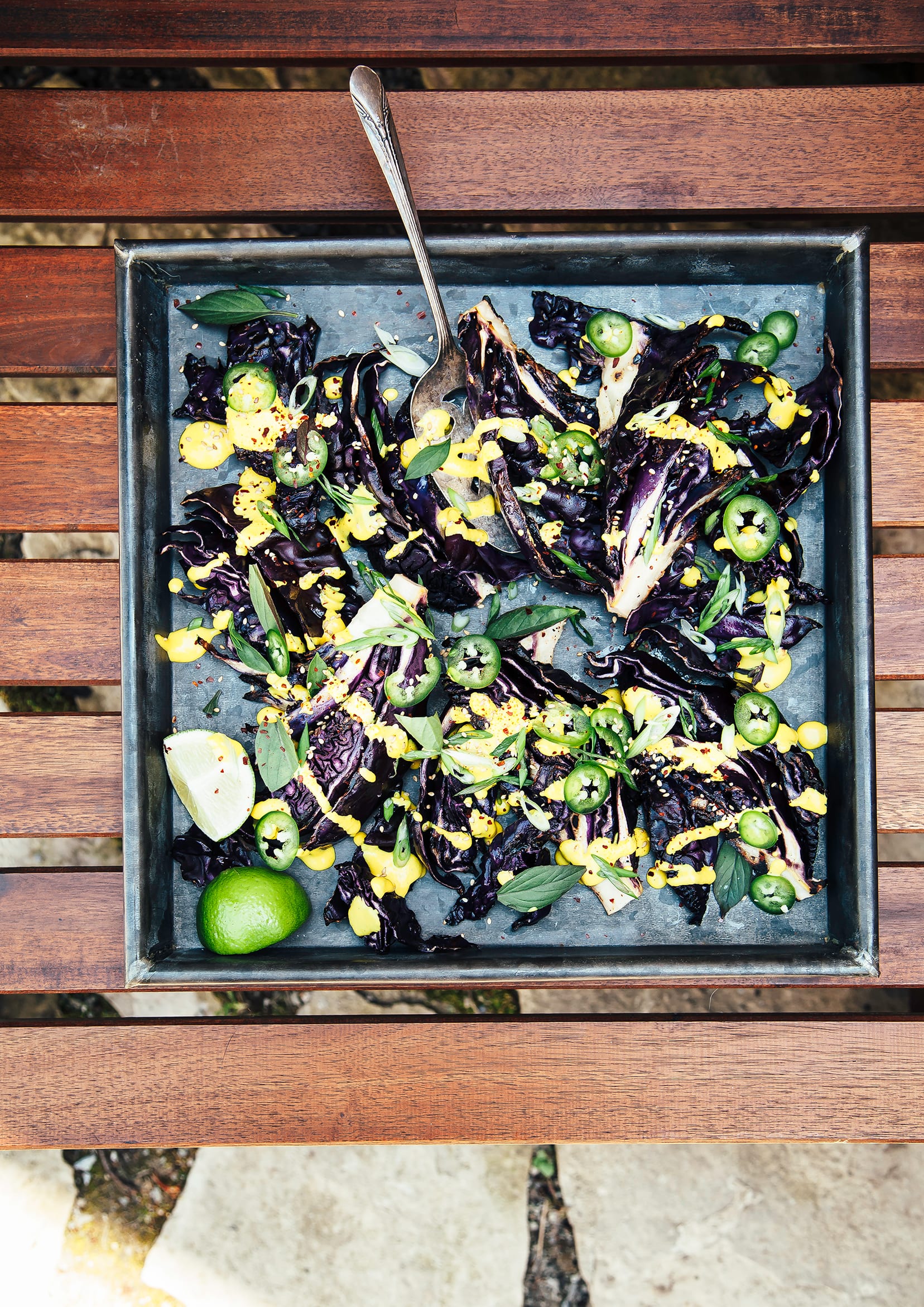 Image shows a dark metal tray with grilled cabbage wedges, drizzled in a creamy bright yellow sauce and topped with chopped herbs and sliced jalapeños .