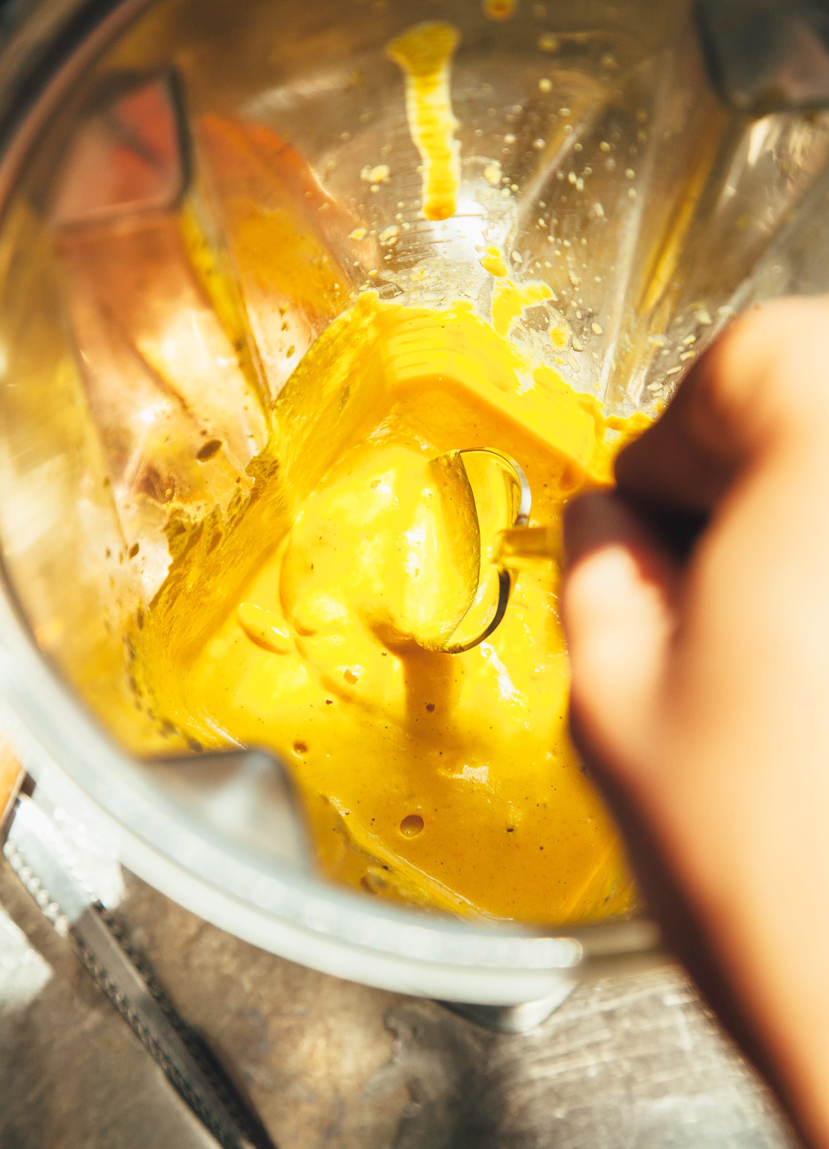 Image shows hand dipping a spoon into a blender of creamy, yellow sauce.