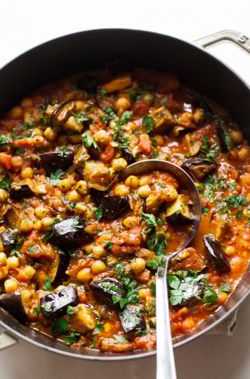 An overhead shot of a stew in a braiser-style pot, containing: seared chunks of eggplant, chickpeas, crushed tomatoes, spices, and chopped parsley.