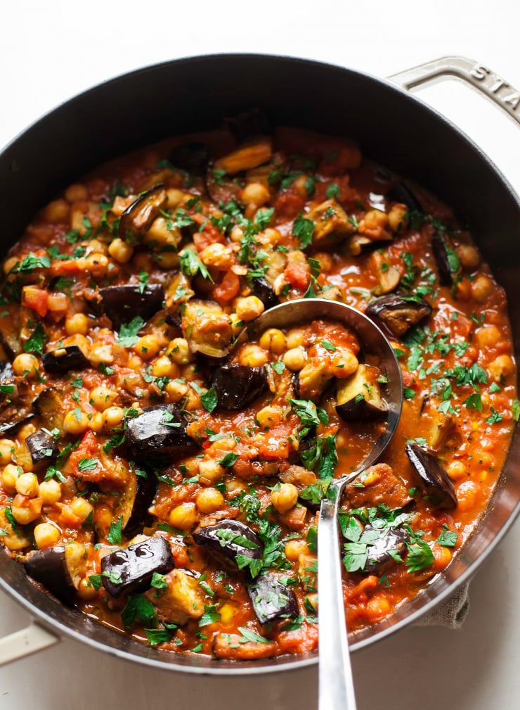 An overhead shot of a light red, chunky stew in a braiser style pot. The stew has chickpeas, chunks of eggplant, and chopped herbs on top.