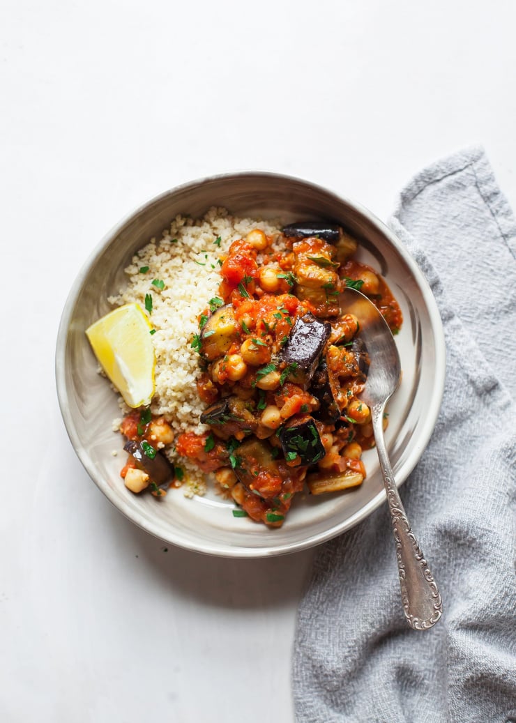 An overhead shot shows an individual serving of braised harissa eggplant with chickpeas in a wide bowl. Alongside is a pile of fluffy cooked millet.
