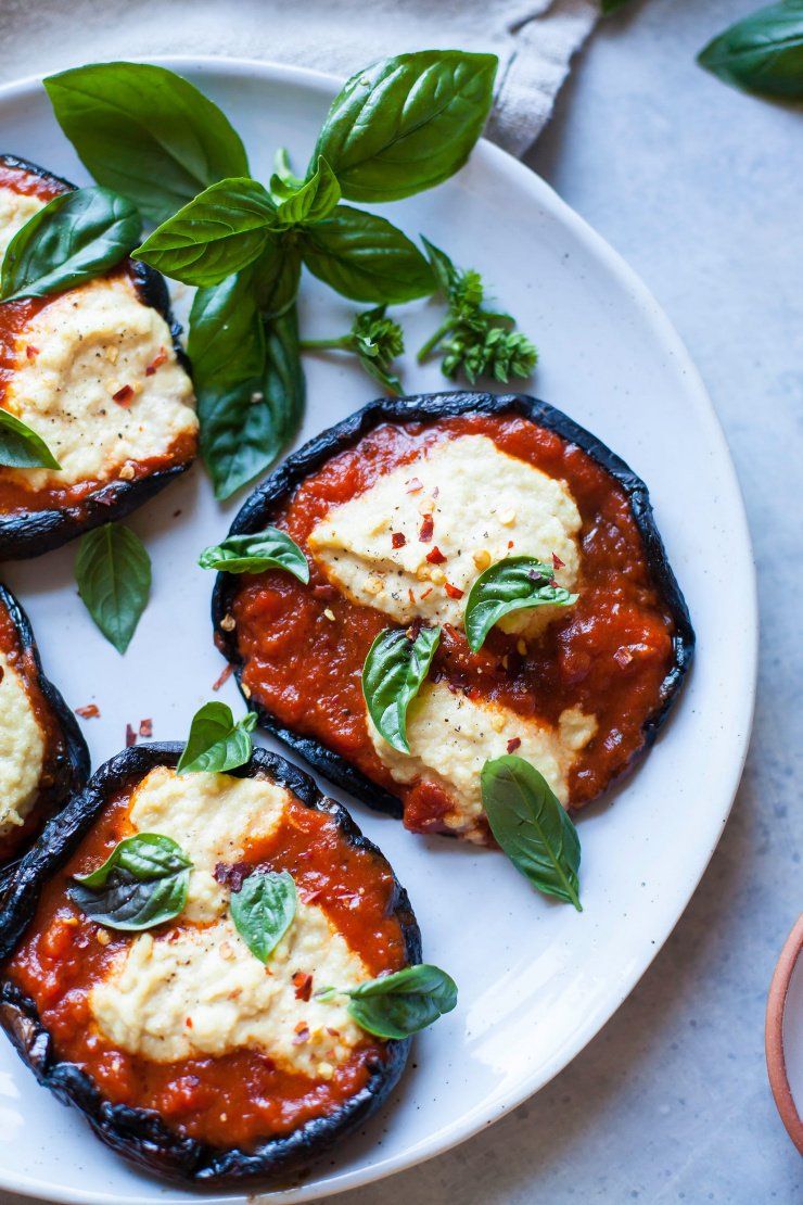 An overhead shot of grilled portobello mushroom caps stuffed with marinara sauce and a creamy cashew-based  "ricotta" mixture. The portobellos are topped with fresh basil leaves.