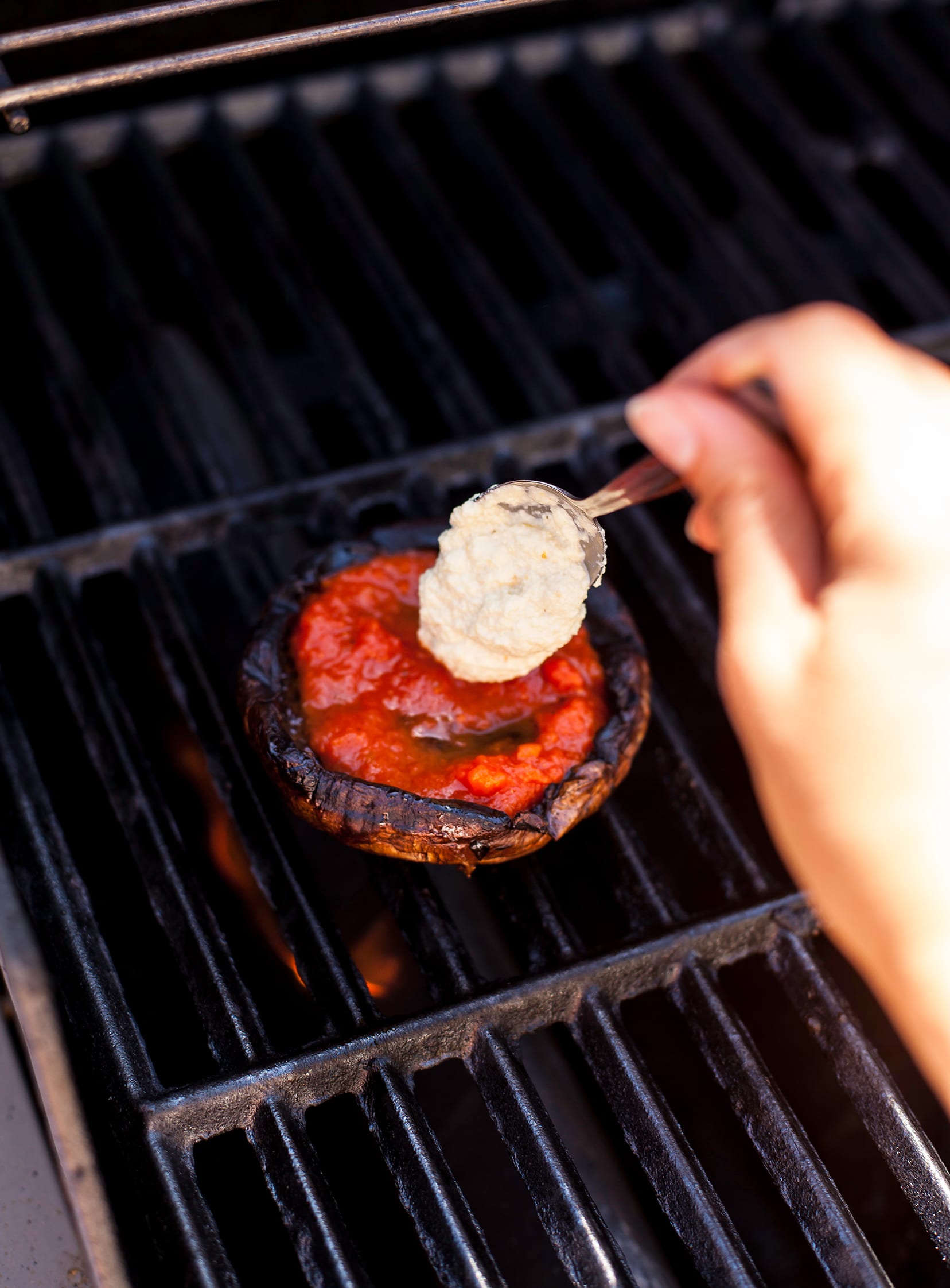 Image shows a hand dollop in lemony cashew “ricotta” into a grilled portobello mushroom cap while it is still on the grill.