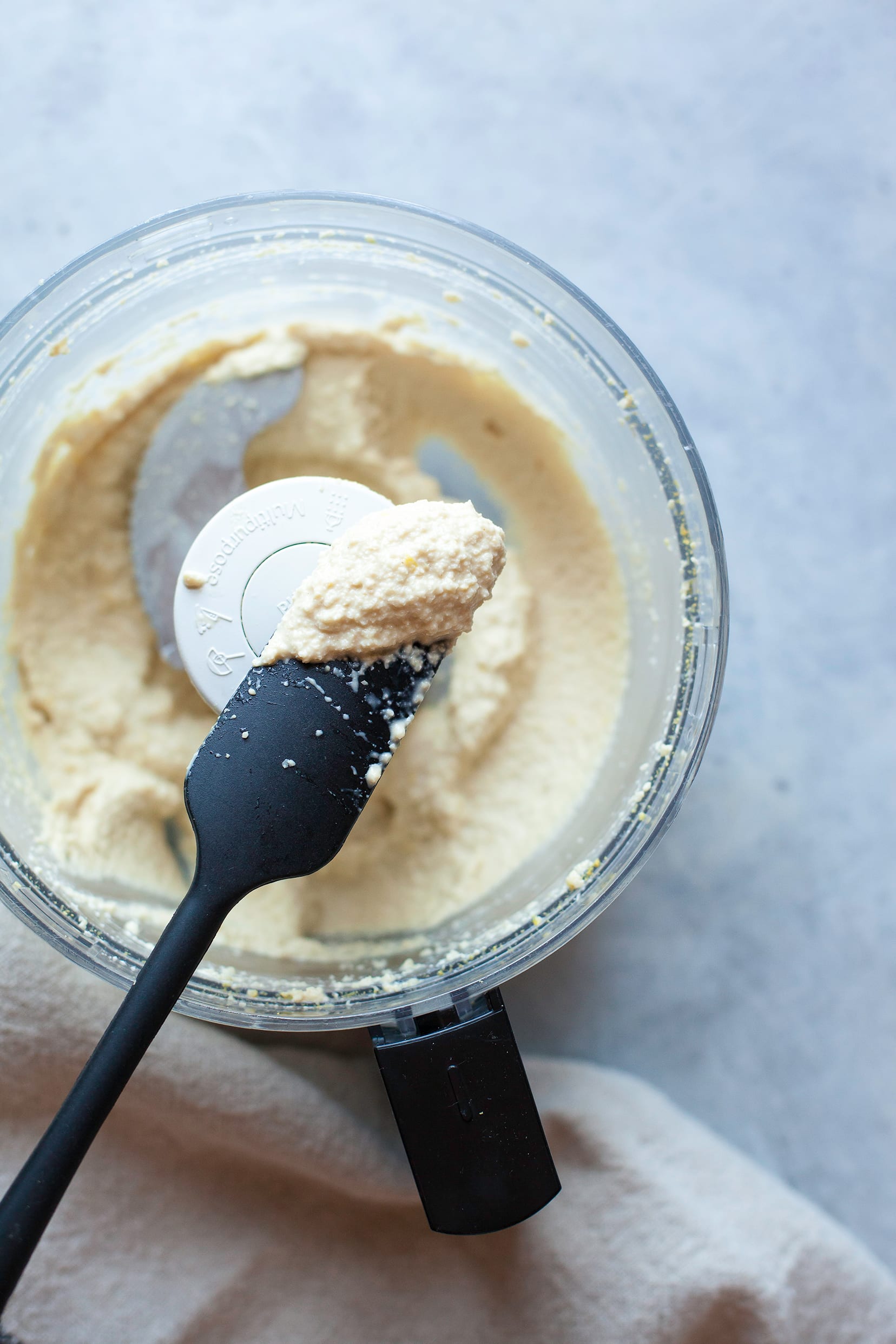 Overhead shot of whipped lemony cashew “ricotta” in the bowl of a food processor with a black spatula.