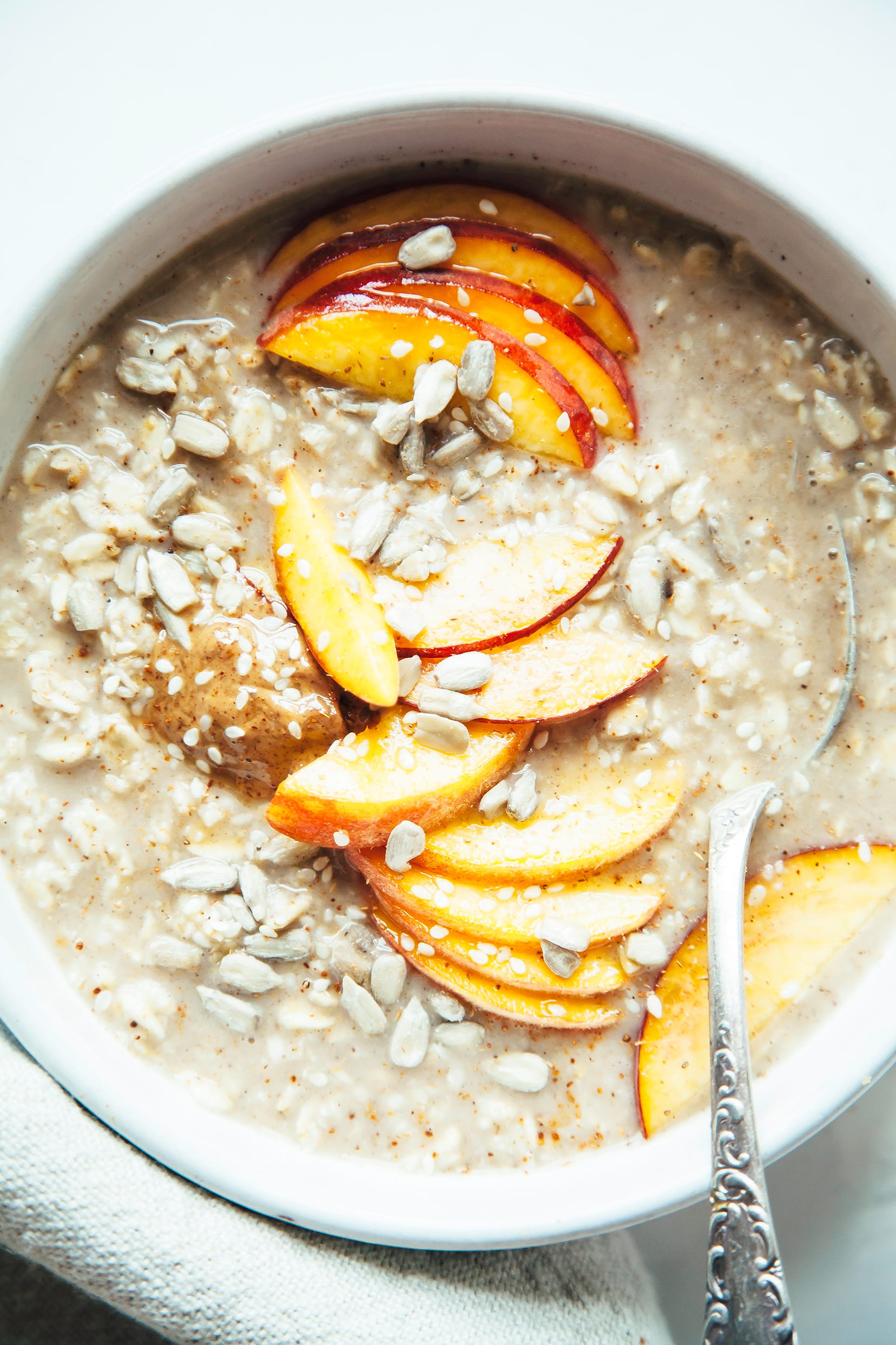 An overhead shot of a bowl of oatmeal, topped with sliced peaches, sunflower seeds, and a dollop of almond butter.