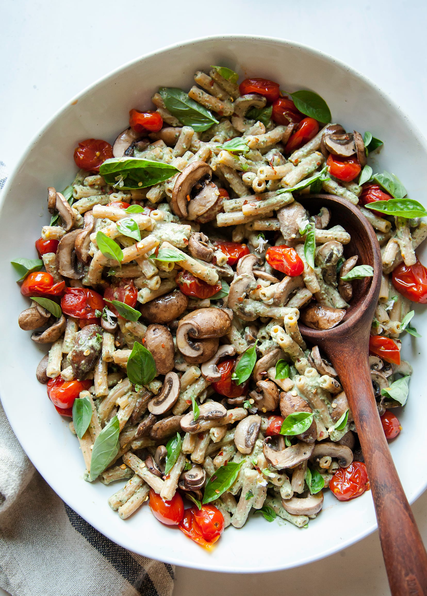 An overhead shot of a creamy basil tahini pasta with burst tomatoes and mushrooms. The pasta is in a large white bowl with a wooden serving spoon.