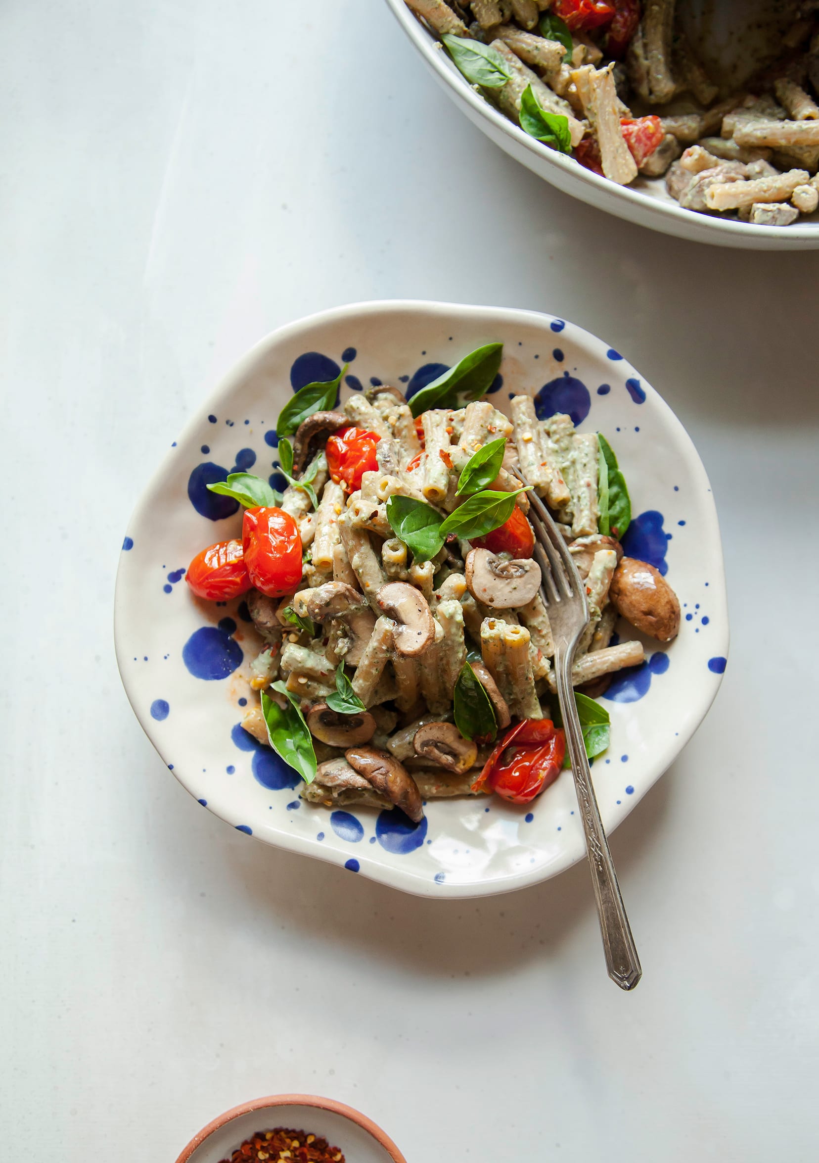 An overhead image of a basil tahini pasta with burst tomatoes and mushrooms in a blue speckled bowl.