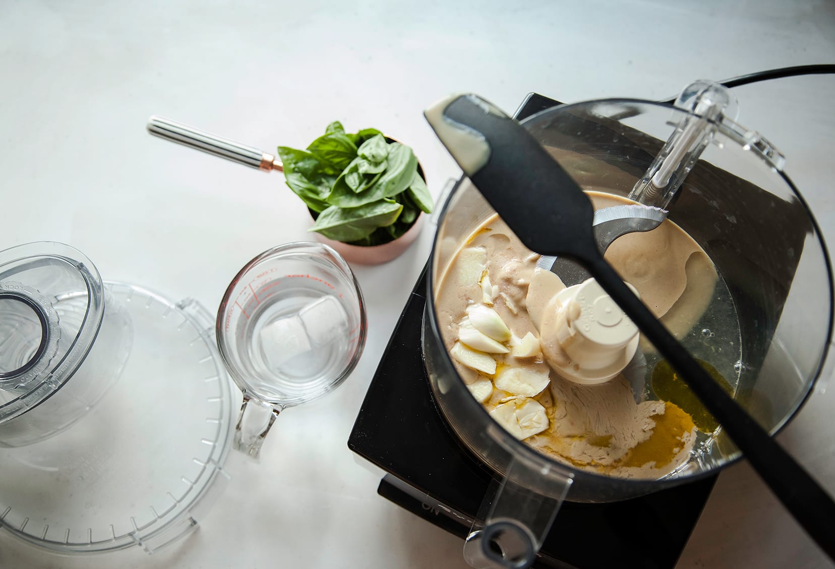 An overhead image of a food processor bowl with tahini and garlic inside. Basil in shown nearby in a measuring cup.