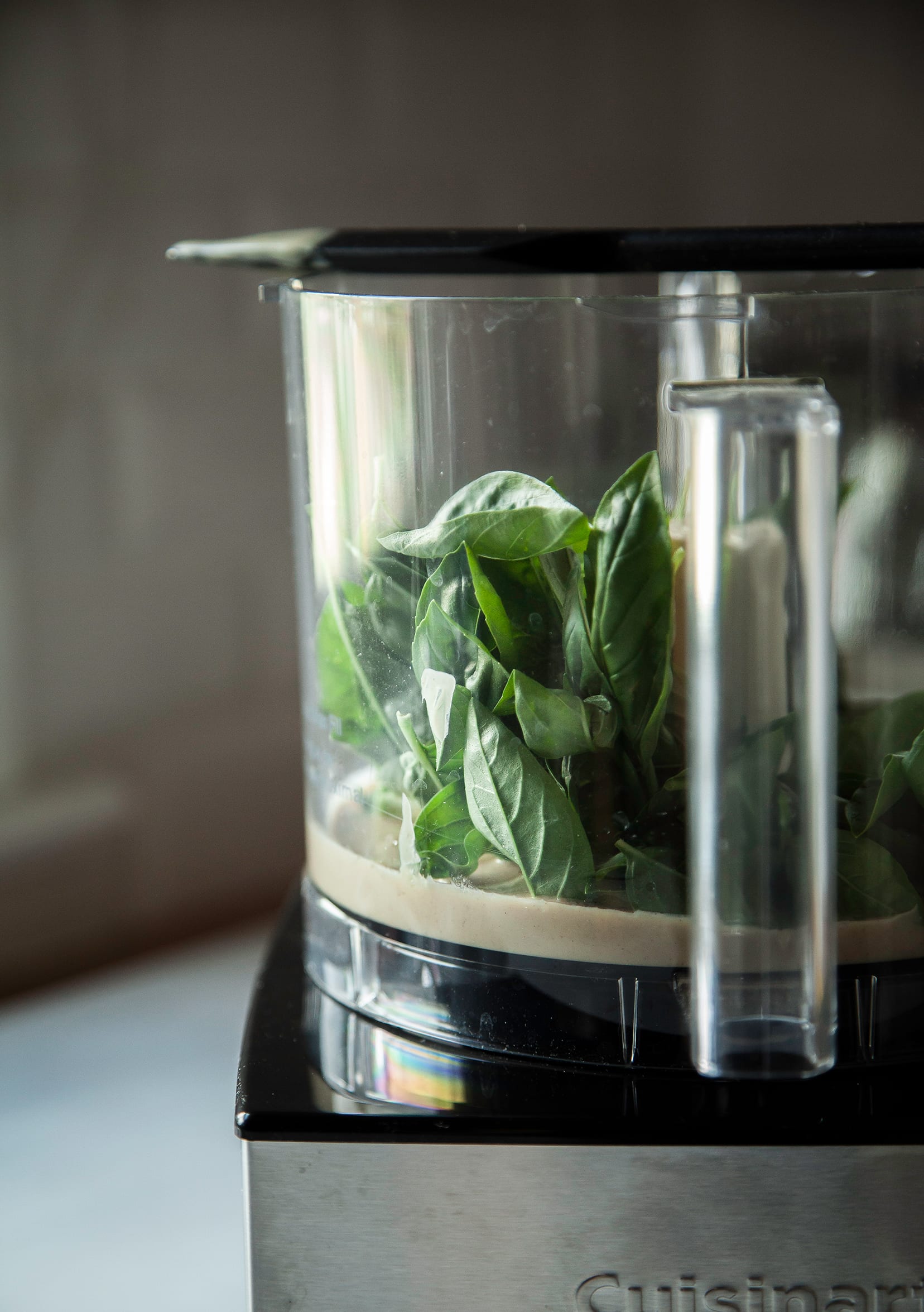 A head-on shot of a food processor bowl on the base with tahini and a bunch of basil leaves within.