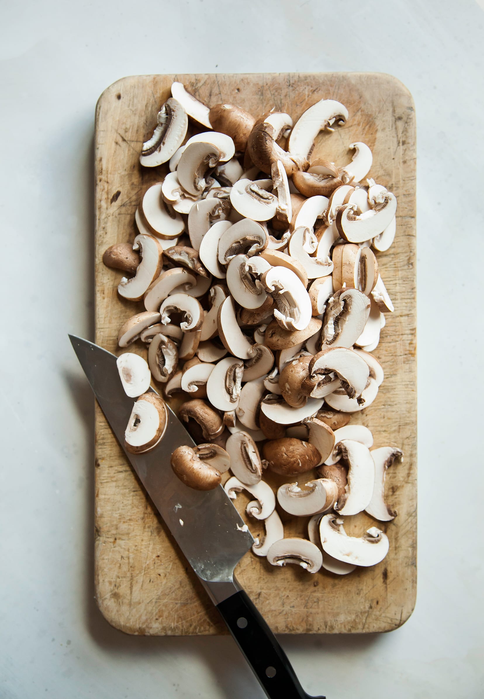 Image shows a bunch of sliced core mini mushrooms on a wooden cutting board with a chef knife to the side.