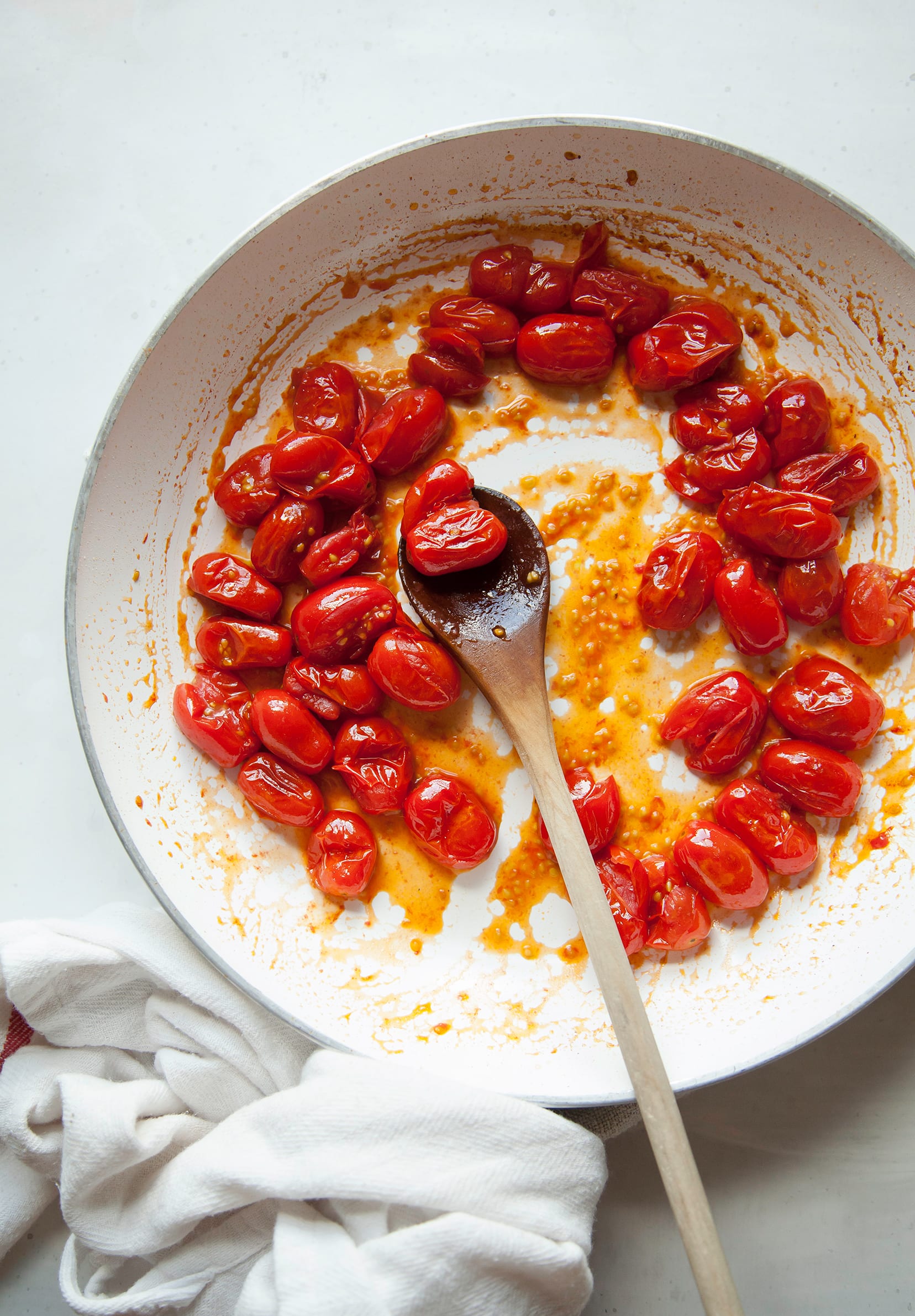 Image shows grape tomatoes being cooked in a white sauté pan with a wooden spoon sticking out of the pan.