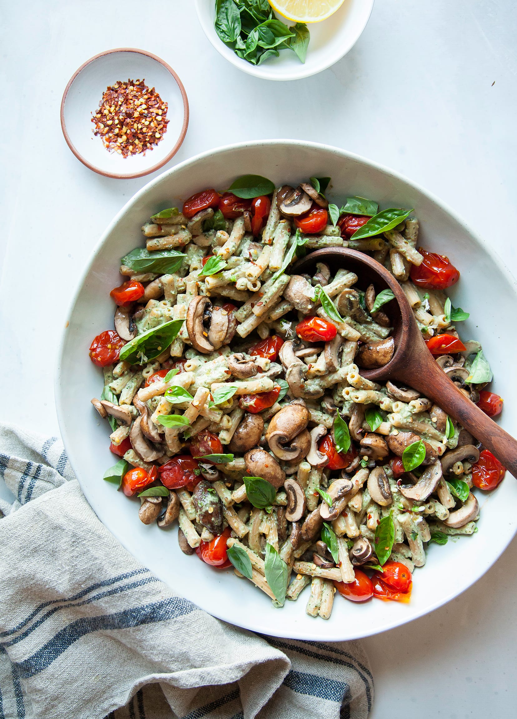 An overhead shot of a creamy basil tahini pasta with burst tomatoes and mushrooms. The pasta is in a large white bowl with a wooden serving spoon.