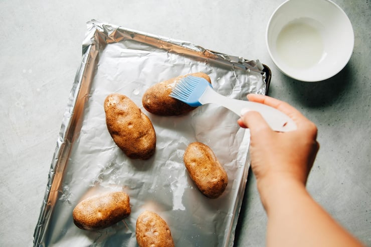 Image shows a hand brushing russet potatoes with some oil.