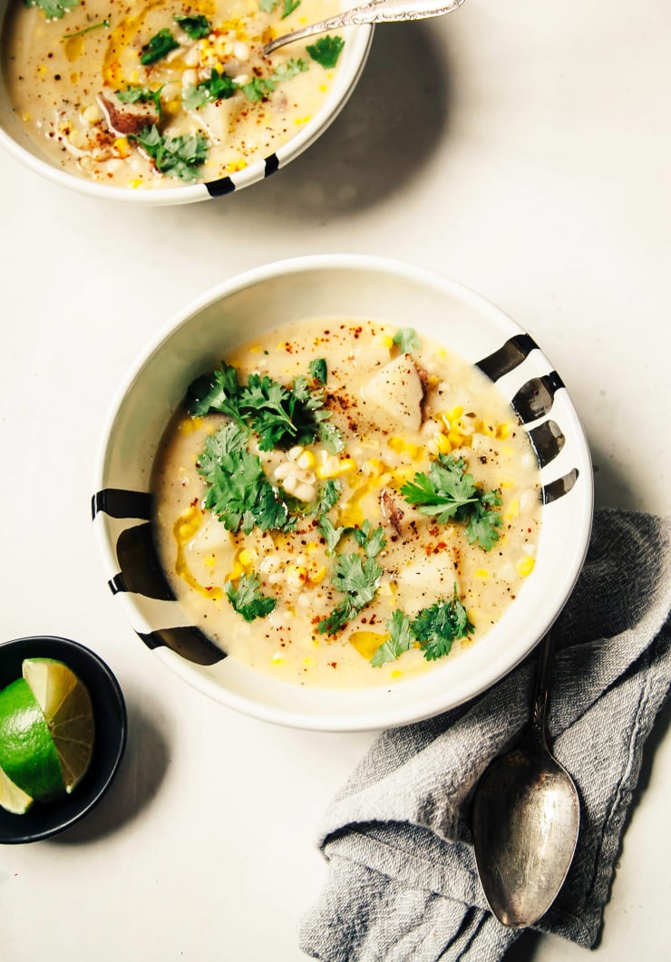An overhead shot of a creamy vegan corn chowder in two white bowls with black streaks, all on a white background. The chowder is pale, golden yellow and garnished with chopped cilantro.