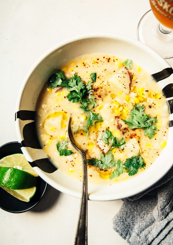 An overhead shot of a bowl of vegan corn chowder in a white bowl on a white background. A beer is poured nearby and there is a small bowl with lime wedges nearby as well.
