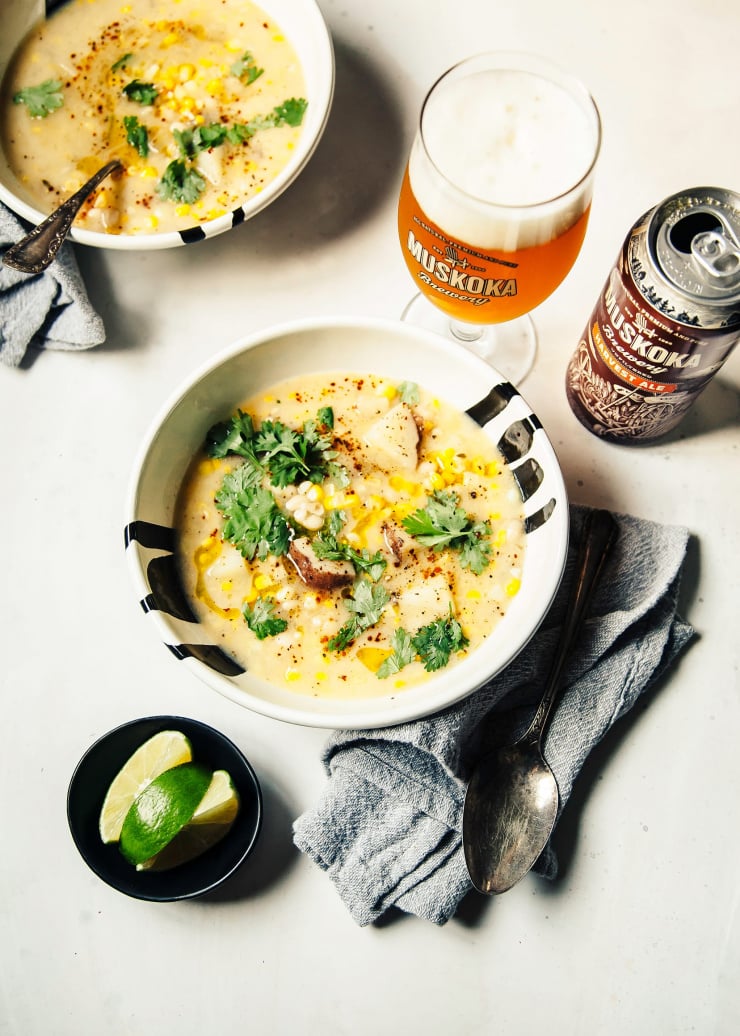 An overhead shot of two bowls of vegan corn chowder in white bowls on a white background. A beer is poured nearby and there is a small bowl with lime wedges nearby as well.
