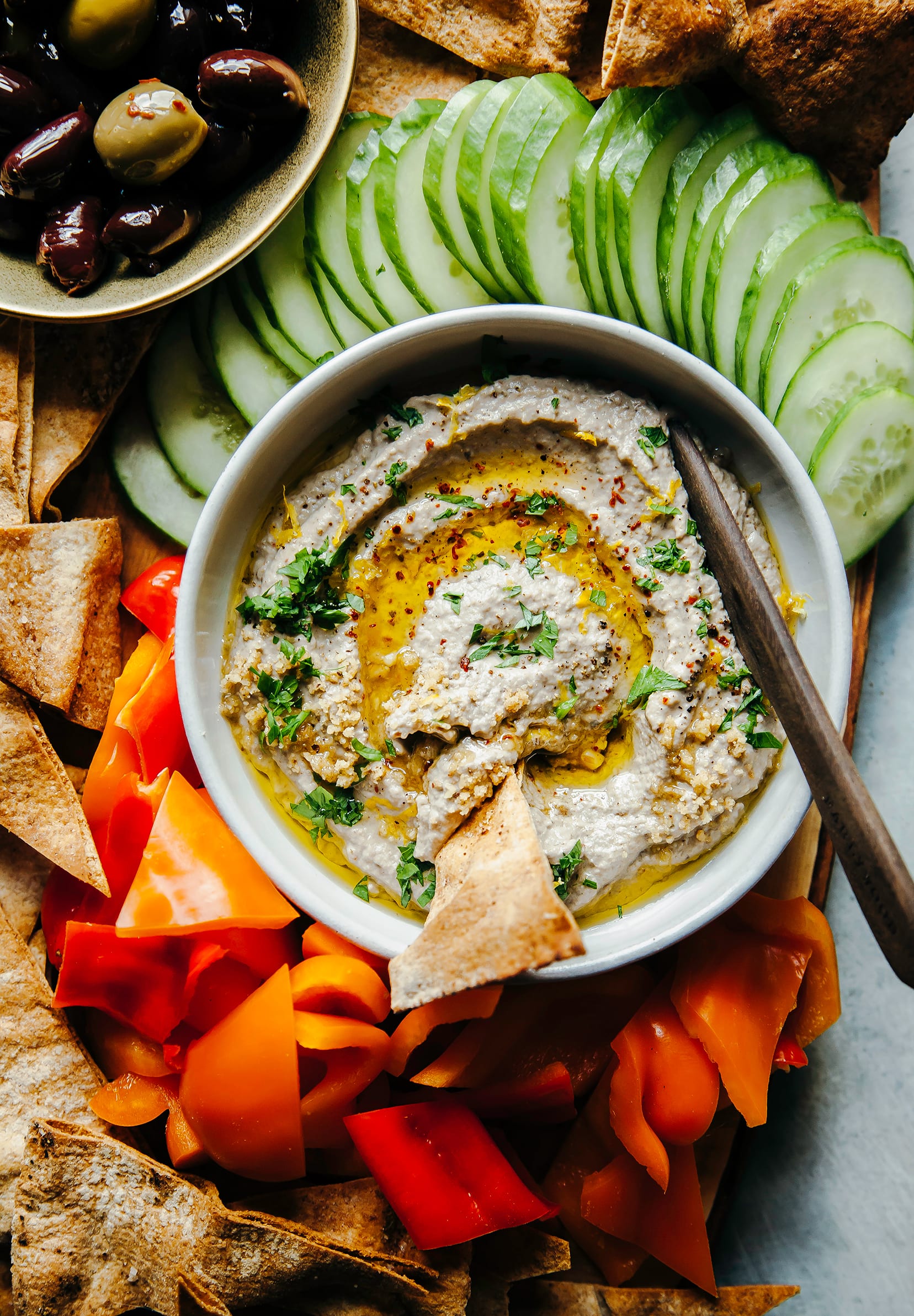 Image shows a board with a bowl of smooth eggplant dip, chopped vegetables, pita chips and olives.