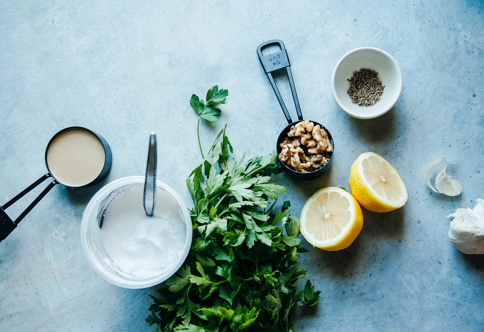 An overhead shot of eggplant dip ingredients: non-dairy yogurt, parsley, lemon, walnuts,  cumin, tahini and garlic.
