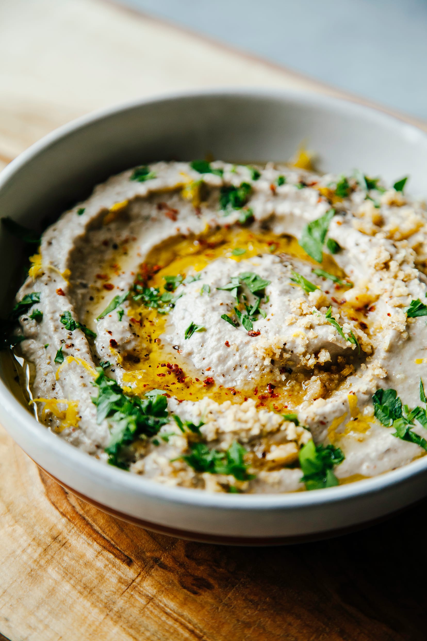 A 3/4 angle shot of a creamy eggplant dip in a white bowl, topped with chopped walnuts, parsley, and olive oil.